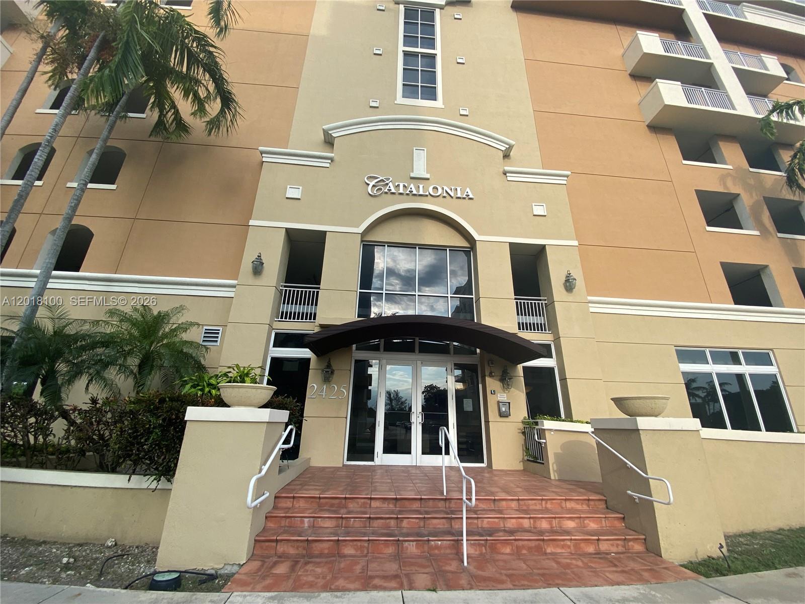 The exterior entrance of a multi-story tan building features a tiled staircase and glass doors — 2425 SW 27th Ave # 1008, Miami FL 33145, MIAMI, FL