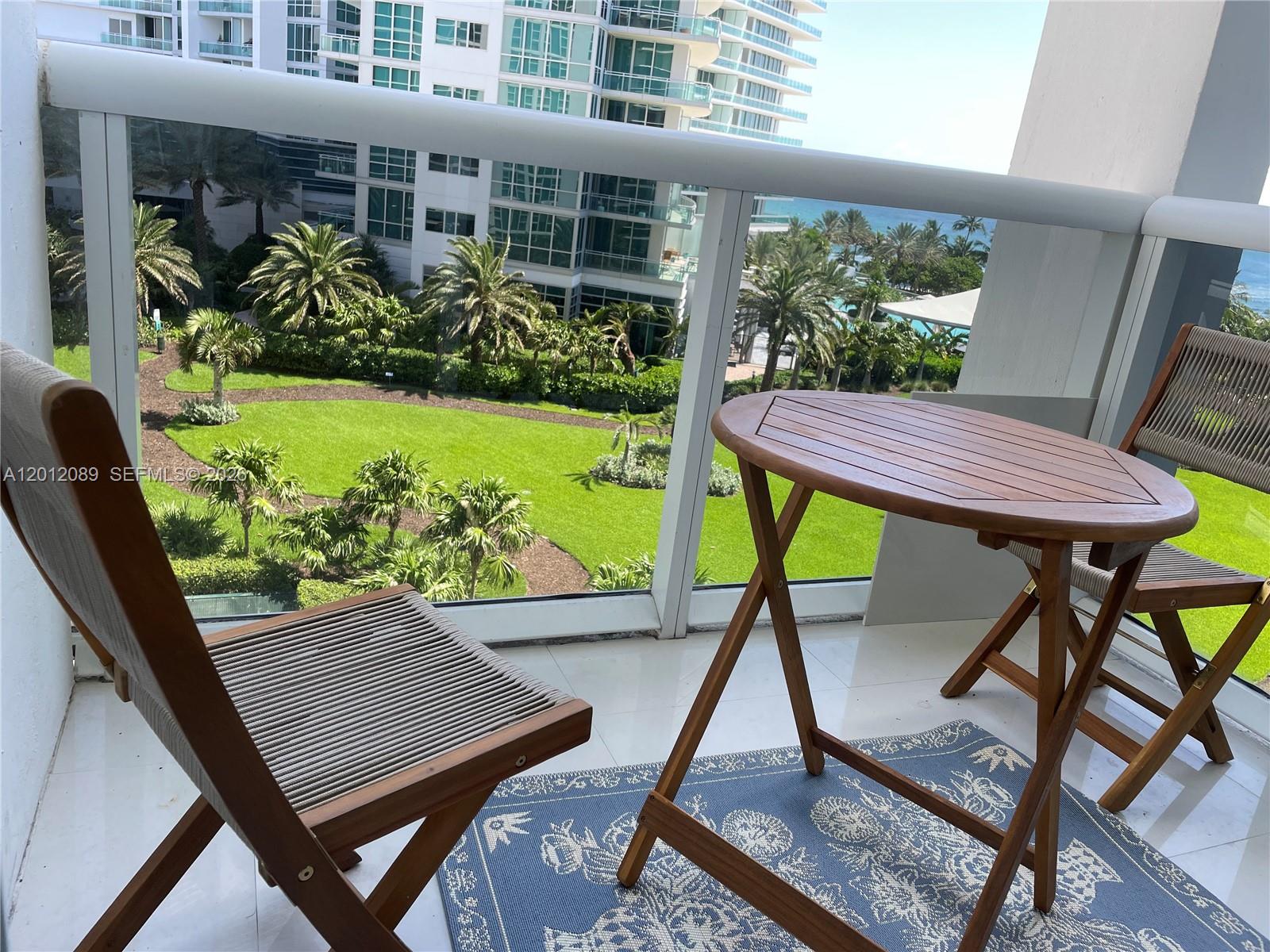 A balcony features a wooden table and two chairs overlooking a landscaped courtyard and ocean — 10275 Collins Ave # 602, Bal Harbour FL 33154, BAL HARBOUR, FL