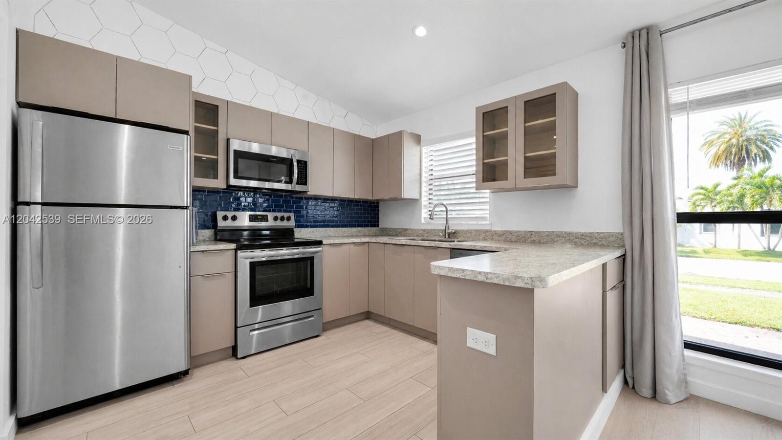 A kitchen features stainless steel appliances, taupe cabinetry, a blue tiled backsplash, and light flooring — 1704 Rodman St # 1, Hollywood FL 33020, HOLLYWOOD, FL