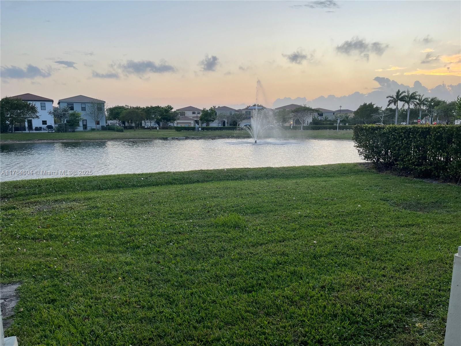 A grassy lawn overlooks a pond with a fountain and houses in the background — 616 NE 193rd St, Miami FL 33179, MIAMI, FL