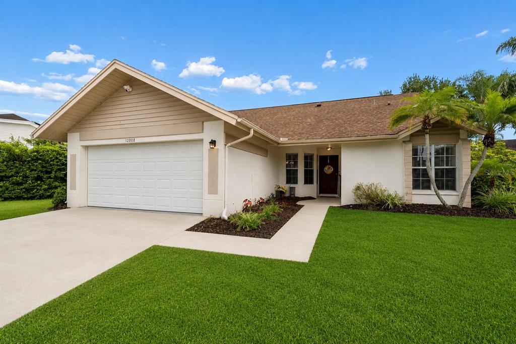 A single-story house with a two-car garage, beige stucco exterior, and a manicured front lawn — 12008 PEONY COURT, TAMPA, FL