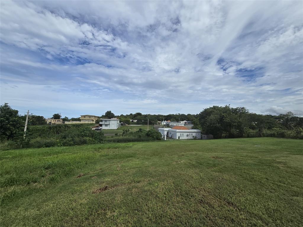 A wide, grassy lot overlooks a residential neighborhood under a cloudy blue sky — BO. GUERRERO MANSIONES DEL CIELO, AGUADILLA, PR