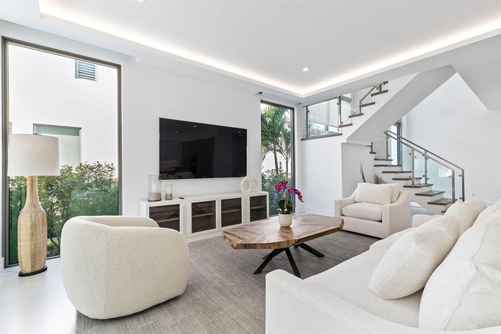 A modern living room features white furniture, a wooden coffee table, and a glass staircase — 406 Ocean One Lane, Juno Beach FL 33408, JUNO BEACH, FL