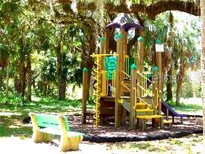 A wooden playground structure with a slide and a bench sits under large oak trees — 24183 SAVORY LANE, PUNTA GORDA, FL