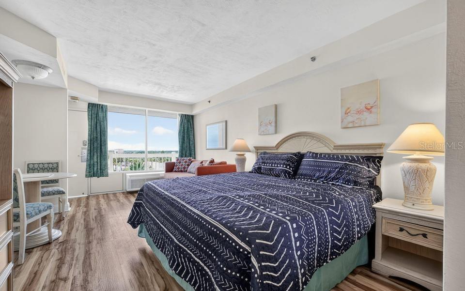 A bedroom features a king bed, wood-look flooring, a small dining set, and balcony access — 2700 N ATLANTIC AVENUE UNIT 507, DAYTONA BEACH, FL