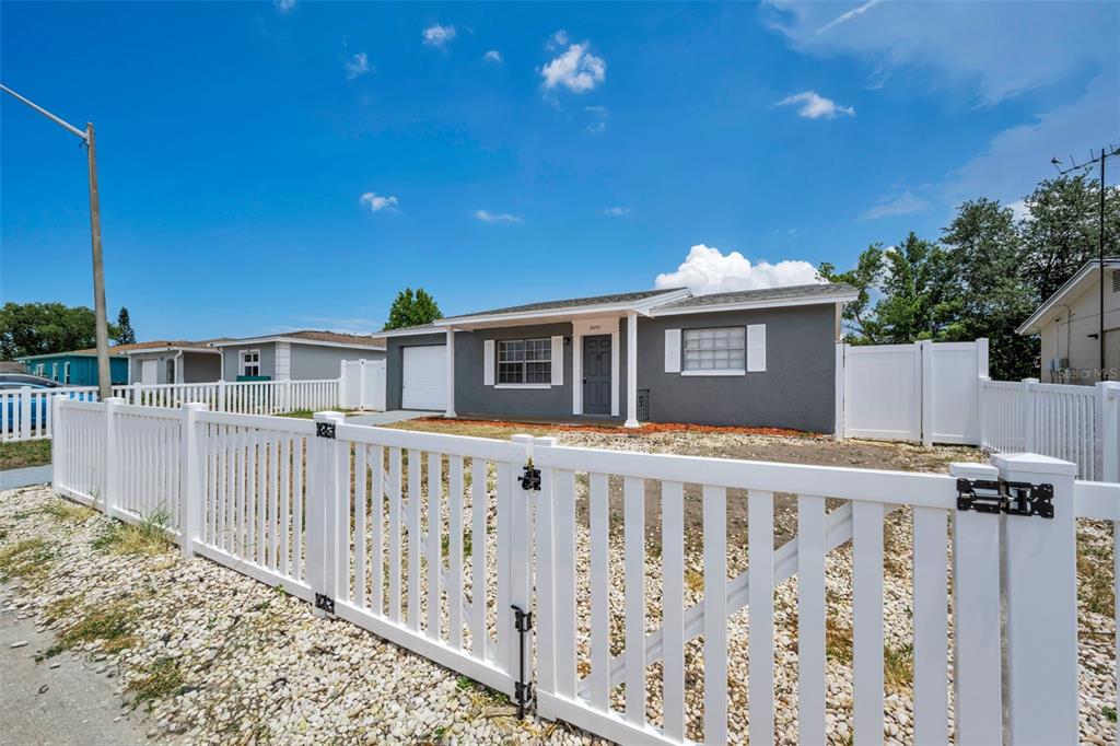 A gray single-story house with a white vinyl fence and gravel yard under a blue sky — 8053 DEDHAM DRIVE, PORT RICHEY, FL