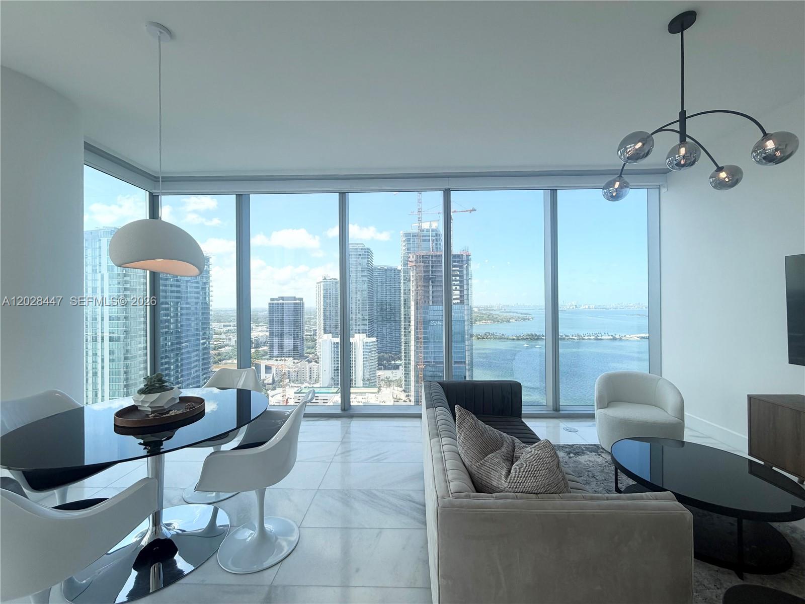 A modern living and dining area features floor-to-ceiling windows overlooking a city skyline and water — 700 NE 26th Ter # 3206, Miami FL 33137, MIAMI, FL