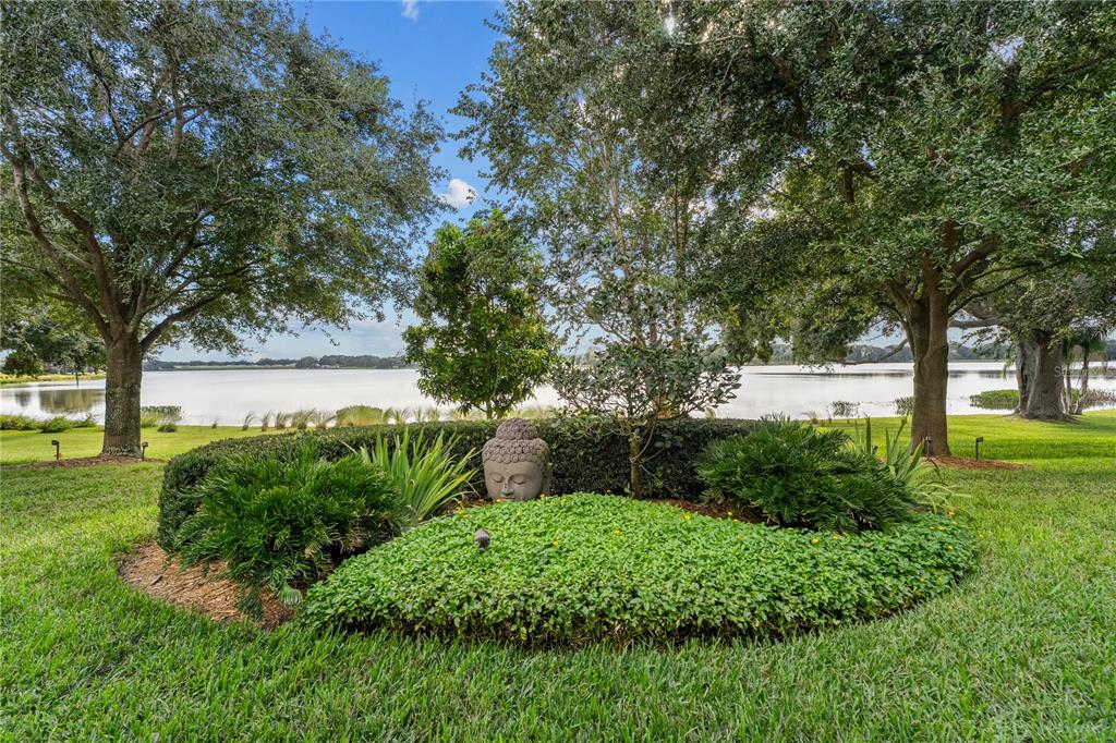A stone Buddha head statue sits in a landscaped garden bed overlooking a calm lake — 414 MEANDERING WAY, POLK CITY, FL