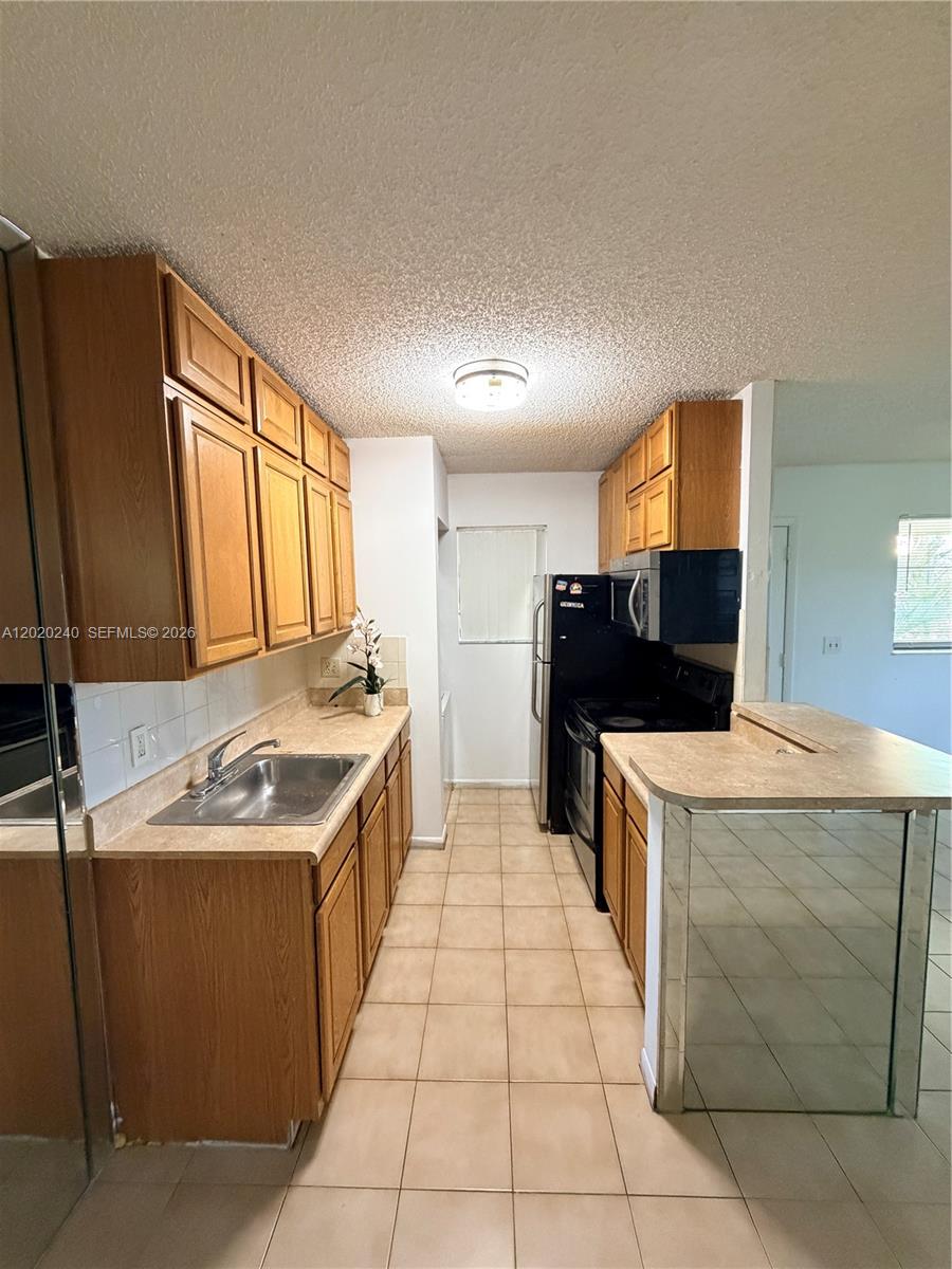 A galley kitchen features light wood cabinets, beige countertops, and black appliances — 588 Trace Cir # 202, Deerfield Beach FL 33441, DEERFIELD BEACH, FL