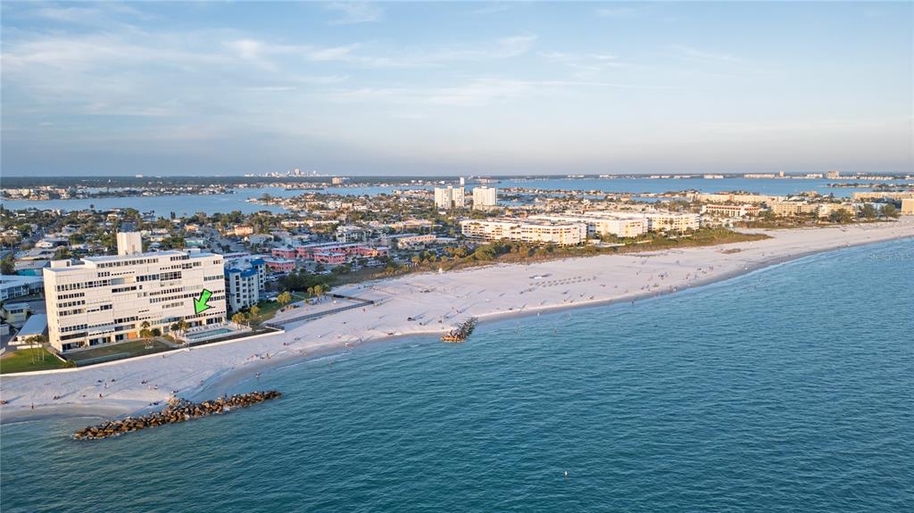 An aerial view shows a white beachfront condominium building with a pool near the ocean — 7000 BEACH PLAZA UNIT 202, ST PETE BEACH, FL