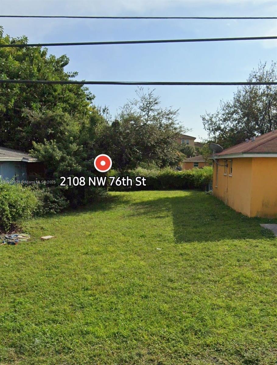 A grassy backyard sits between two residential buildings with dense trees in the background — 2108 NW 76th St, Miami FL 33147, MIAMI, FL