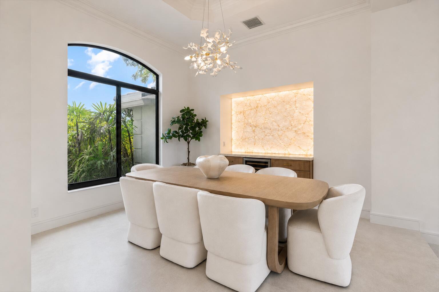 A dining room features a wooden table, white upholstered chairs, a chandelier, and backlit stone — 15463 Tall Oak Avenue, Delray Beach FL 33446, DELRAY BEACH, FL