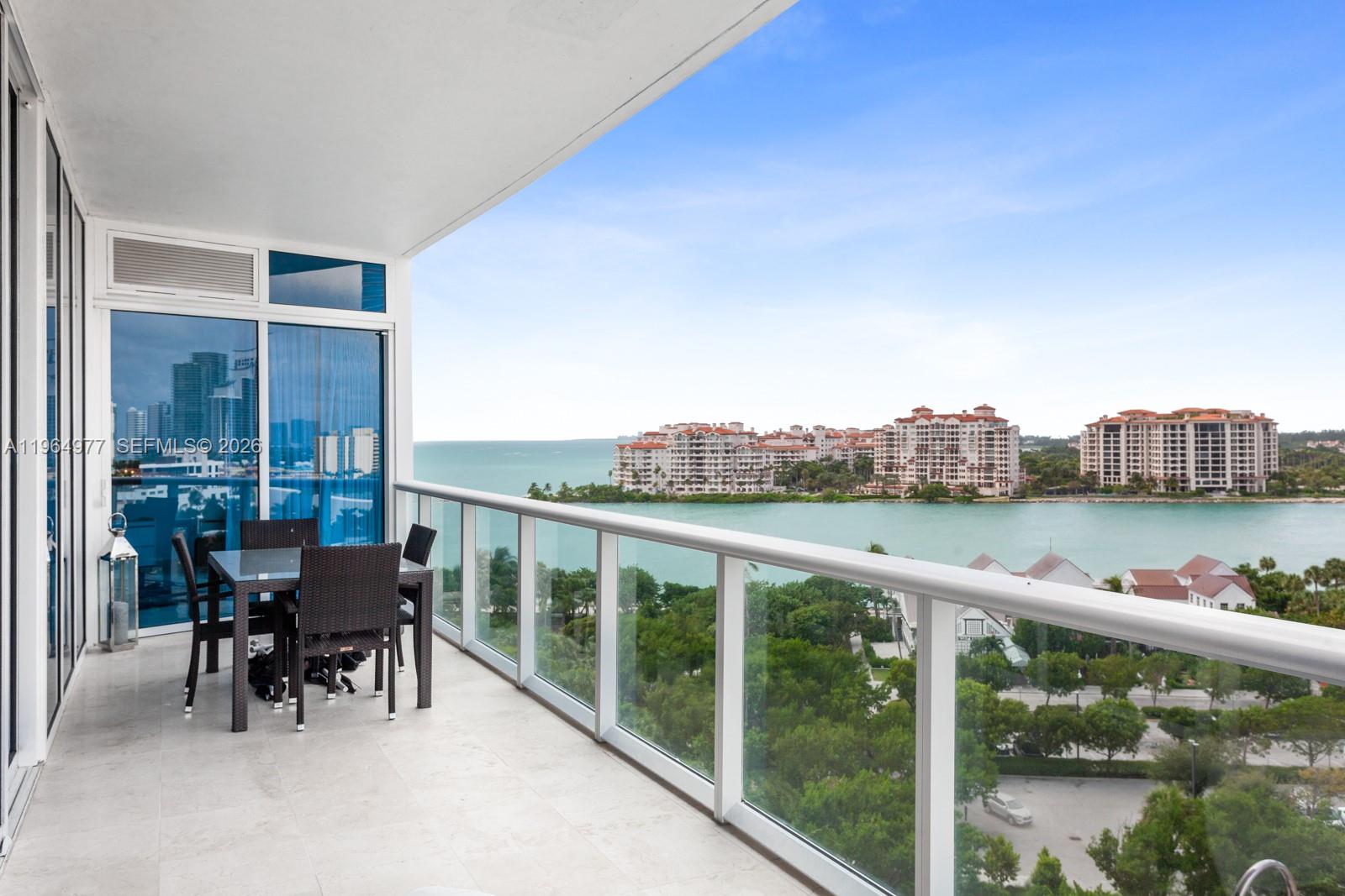 A balcony with a dining set overlooks a bay, lush trees, and distant apartment buildings — 100 S Pointe Dr # 1001, Miami Beach FL 33139, MIAMI BEACH, FL