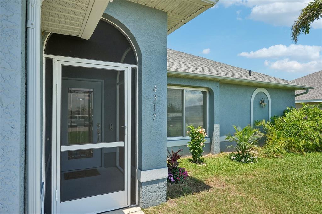 A blue stucco house exterior features an arched entryway with a screen door and landscaping — 4317 NW 40TH TERRACE, CAPE CORAL, FL