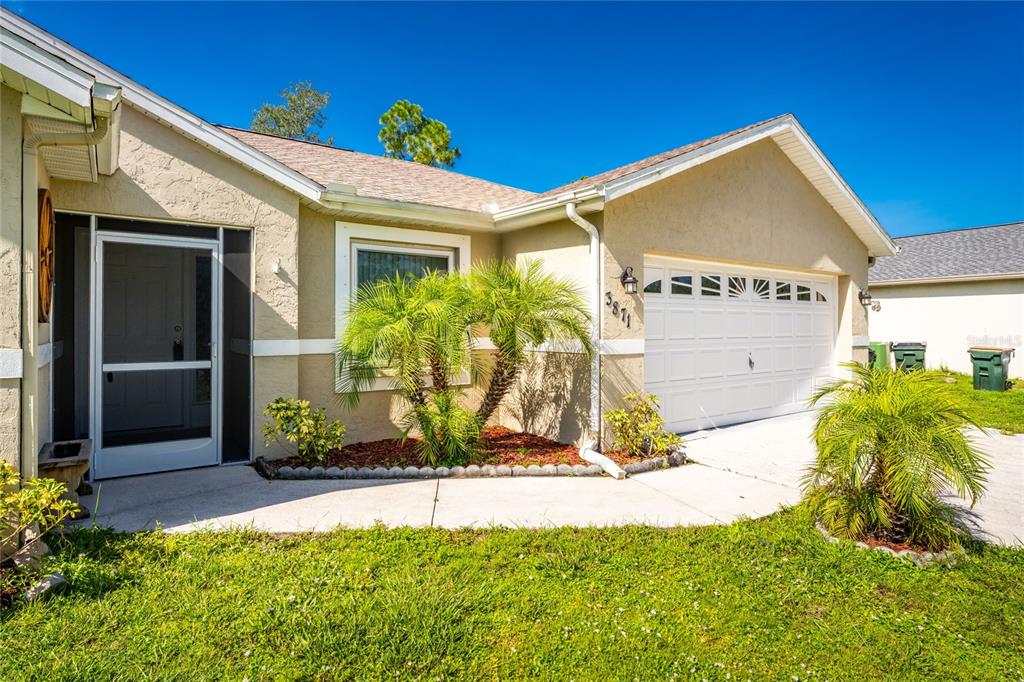 A single-story house exterior features a white garage door, a screened entryway, and landscaping — 3871 COQUINA AVENUE, NORTH PORT, FL