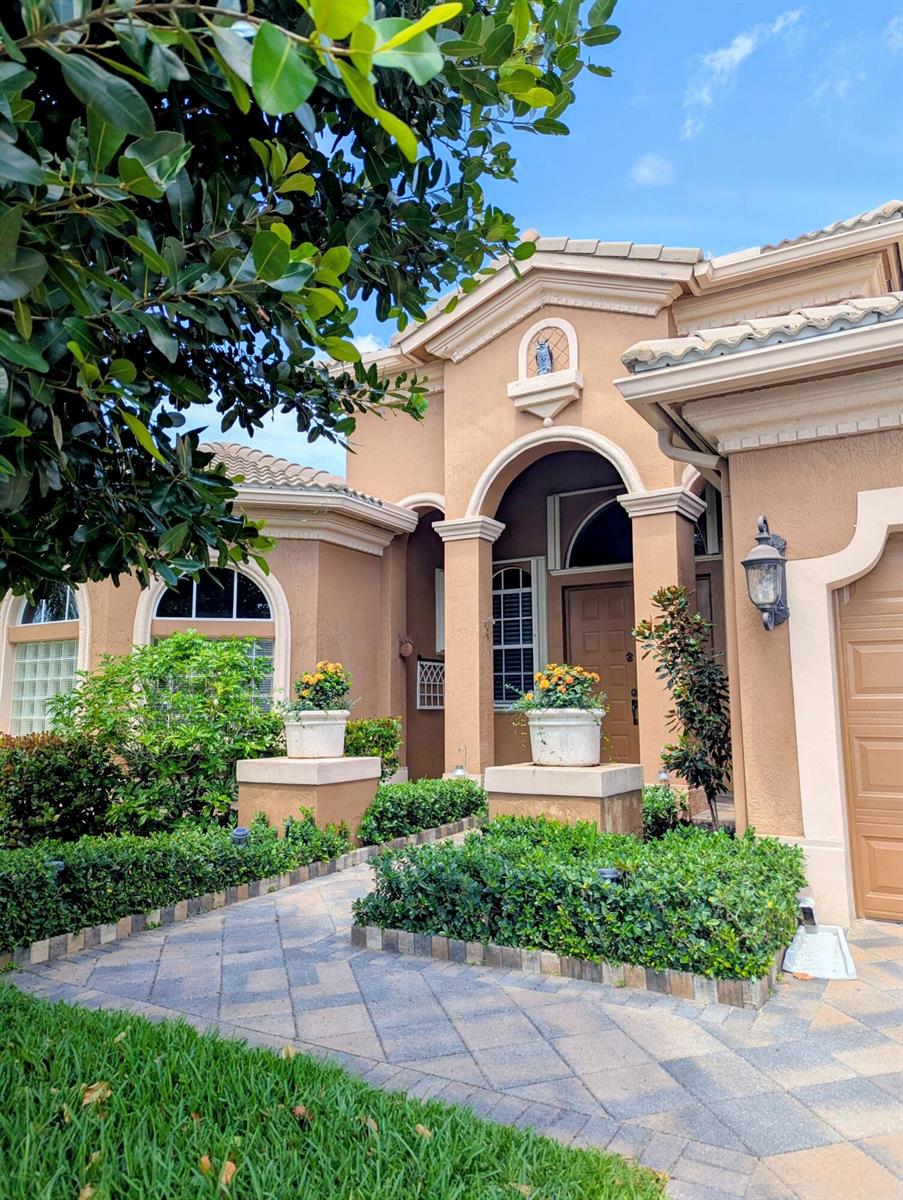 A tan stucco house features a paved walkway, manicured hedges, and two potted flower arrangements — 10640 Oak Meadow Lane, Lake Worth FL 33449, LAKE WORTH, FL