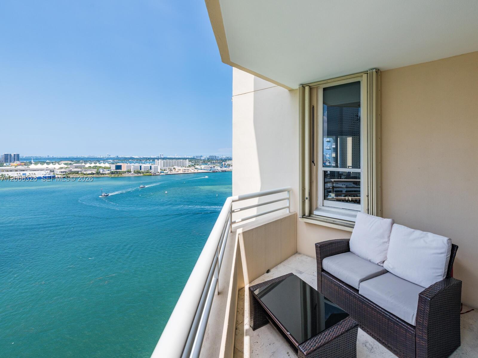 A balcony with wicker furniture overlooks a large body of water and a distant city — 888 Brickell Key Dr # 2112, Miami FL 33131, MIAMI, FL