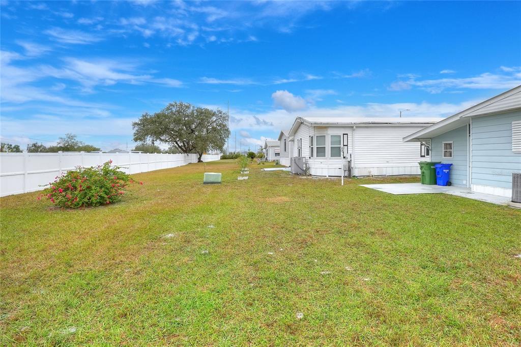 A grassy backyard features a white vinyl fence, a flowering bush, and neighboring mobile homes — 6308 BAYBERRY BOULEVARD NE, WINTER HAVEN, FL