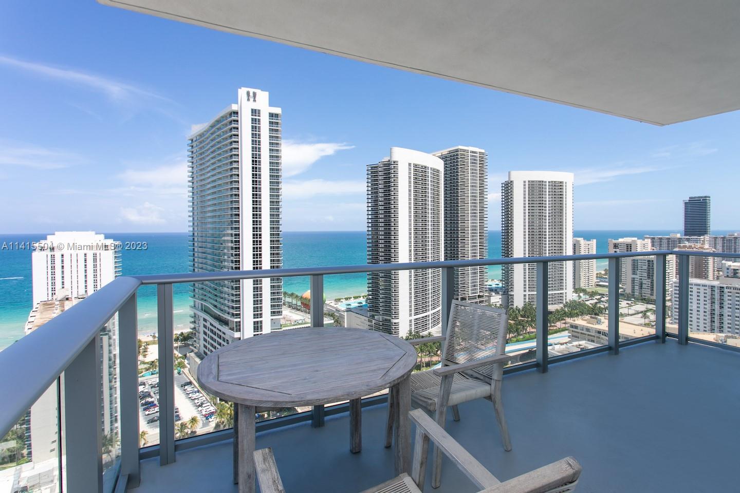A balcony with a wooden table and chairs overlooks the ocean and high-rise buildings — 4010 S Ocean Dr # 1201, Hollywood FL 33019, HOLLYWOOD, FL