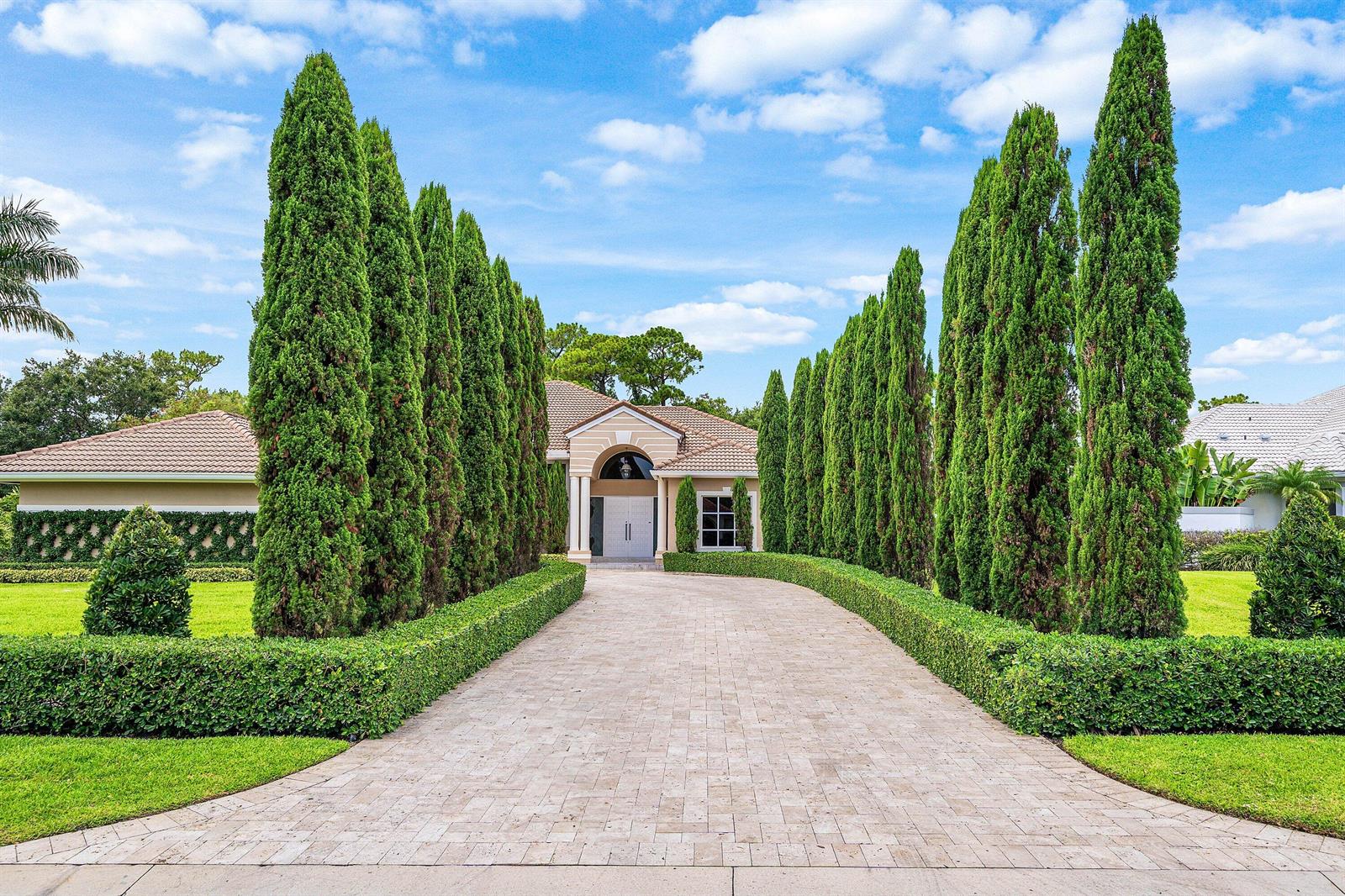 A paved driveway leads to a house entrance framed by tall, manicured cypress trees — 1697 Mayacoo Lakes Boulevard, West Palm Beach FL 33411, WEST PALM BEACH, FL