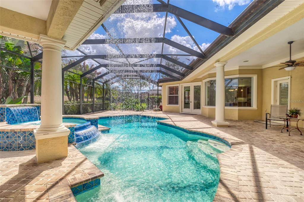 A screened-in pool area features a spa with a waterfall, stone pavers, and white columns — 2983 WENTWORTH WAY, TARPON SPRINGS, FL