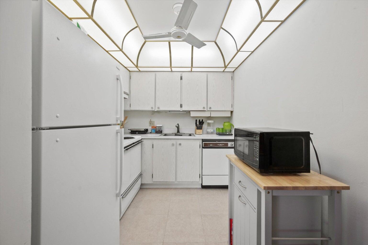 A galley kitchen features white cabinets, white appliances, and a unique arched, backlit ceiling panel — 3300 Springdale Boulevard # 202, Palm Springs FL 33461, PALM SPRINGS, FL