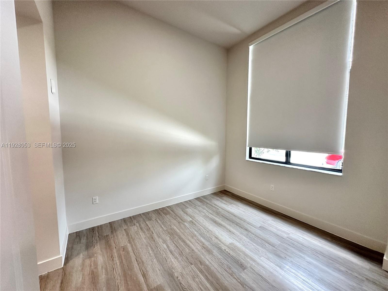An empty room features light-colored walls, wood-look flooring, and a window with a white shade — 18412 Homestead Ave # 110, Miami FL 33157, MIAMI, FL