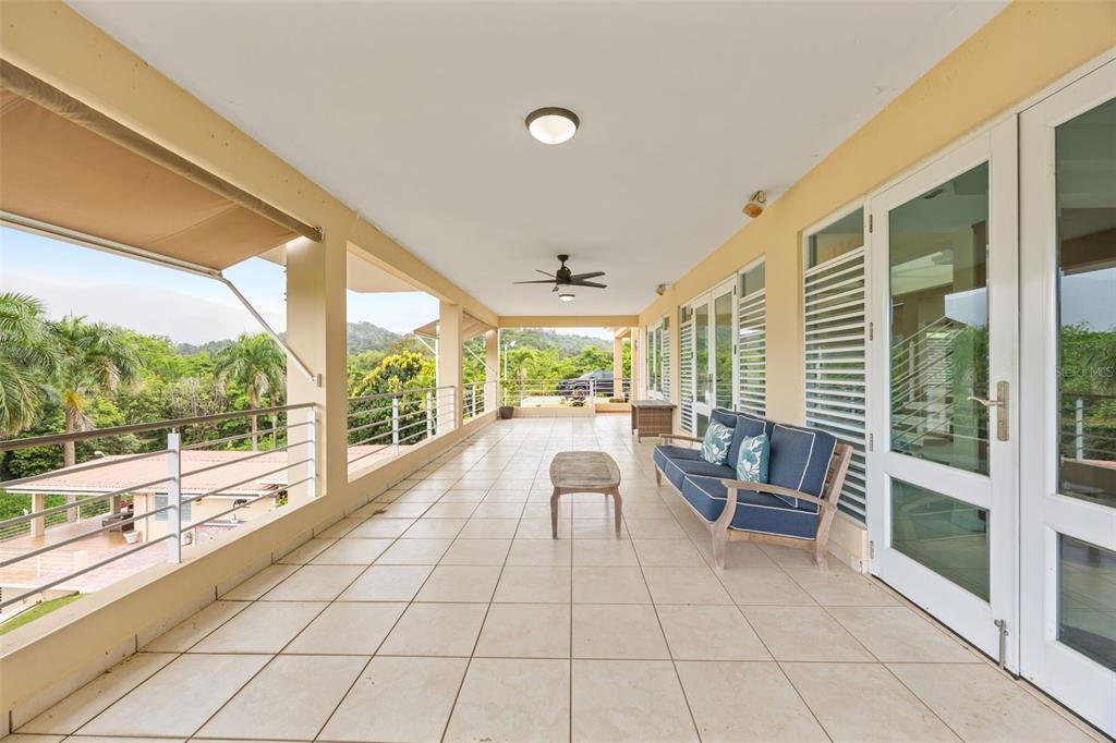 A spacious covered balcony features light tile flooring, a blue sofa, and scenic mountain views — 23 & 24 HACIENDA CARABALI, LUQUILLO, PR