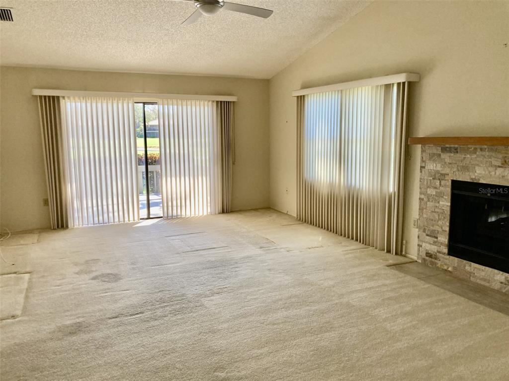 A living room with beige carpet, vertical blinds, a ceiling fan, and a stone fireplace — 3407 AVENIDA MADERA UNIT A, BRADENTON, FL