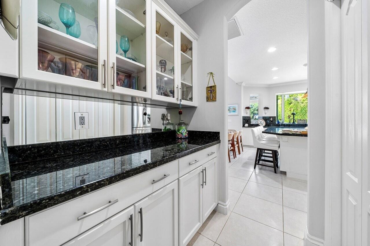 A white kitchen butler's pantry features dark granite countertops, glass-front cabinets, and a mirrored backsplash — 6990 Cypress Cove Circle, Jupiter FL 33458, JUPITER, FL