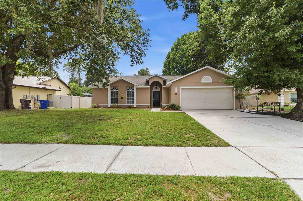 A single-story tan house with a two-car garage and a front lawn under large trees — 1564 CYPRESS WOODS CIRCLE, ST CLOUD, FL