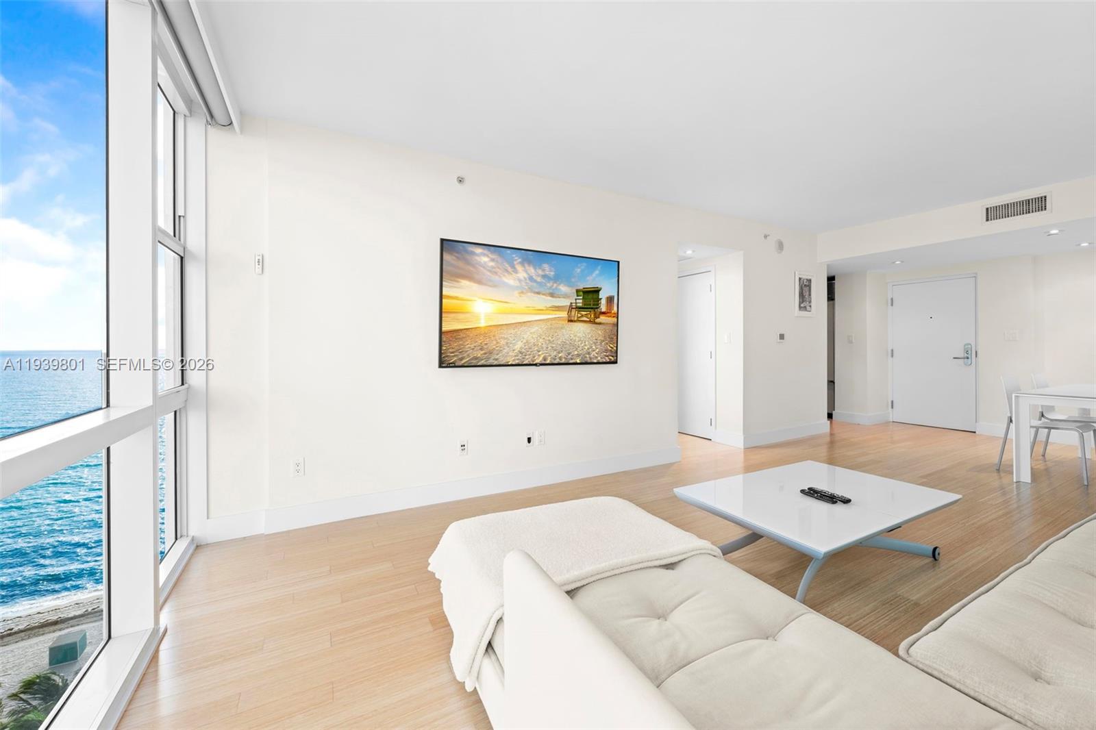 A bright living room features light wood floors, a wall-mounted television, and floor-to-ceiling ocean views — 6801 Collins Ave, Miami Beach FL 33141, MIAMI BEACH, FL