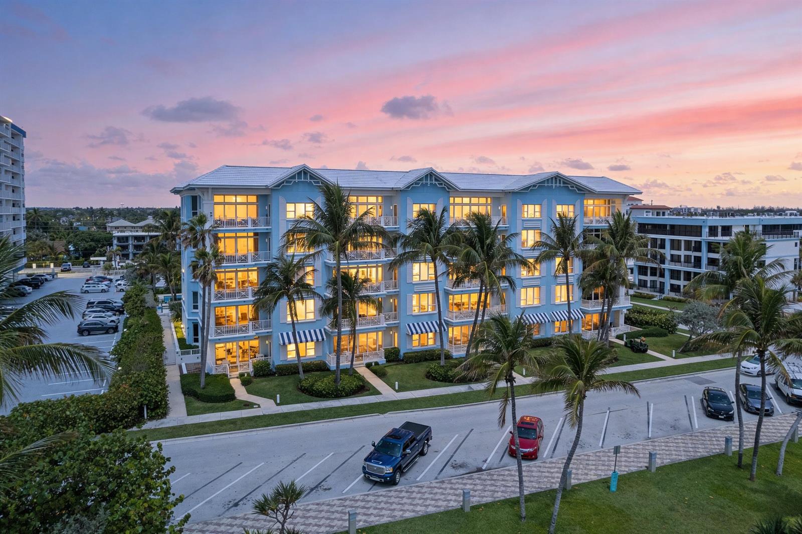 An exterior view of a multi-story blue apartment building at sunset with palm trees — 701 SE 21st Avenue # 109, Deerfield Beach FL 33441, DEERFIELD BEACH, FL