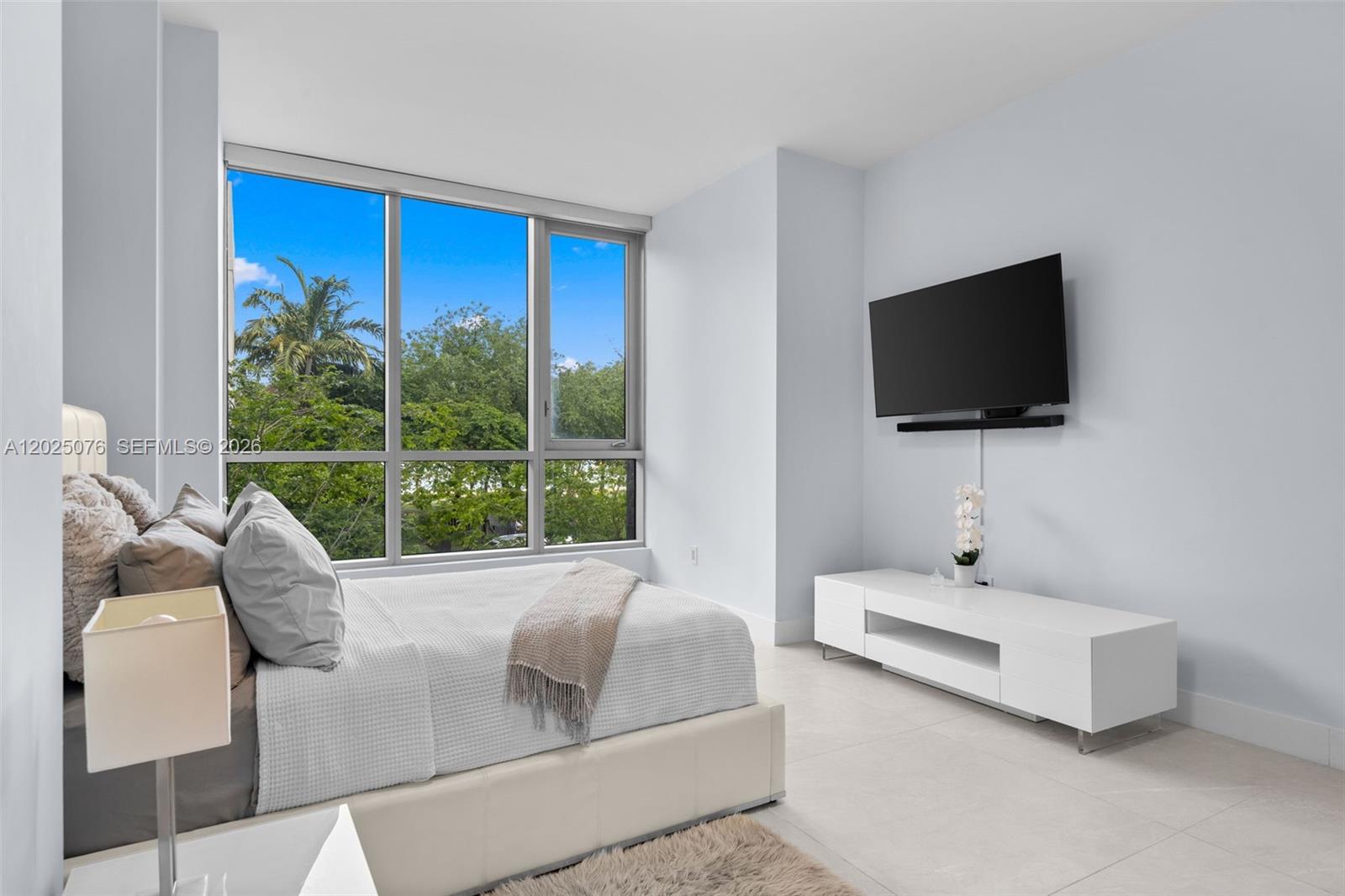 A modern bedroom features a bed, a wall-mounted television, and floor-to-ceiling windows overlooking trees — 650 NE 32nd St, Miami FL 33137, MIAMI, FL