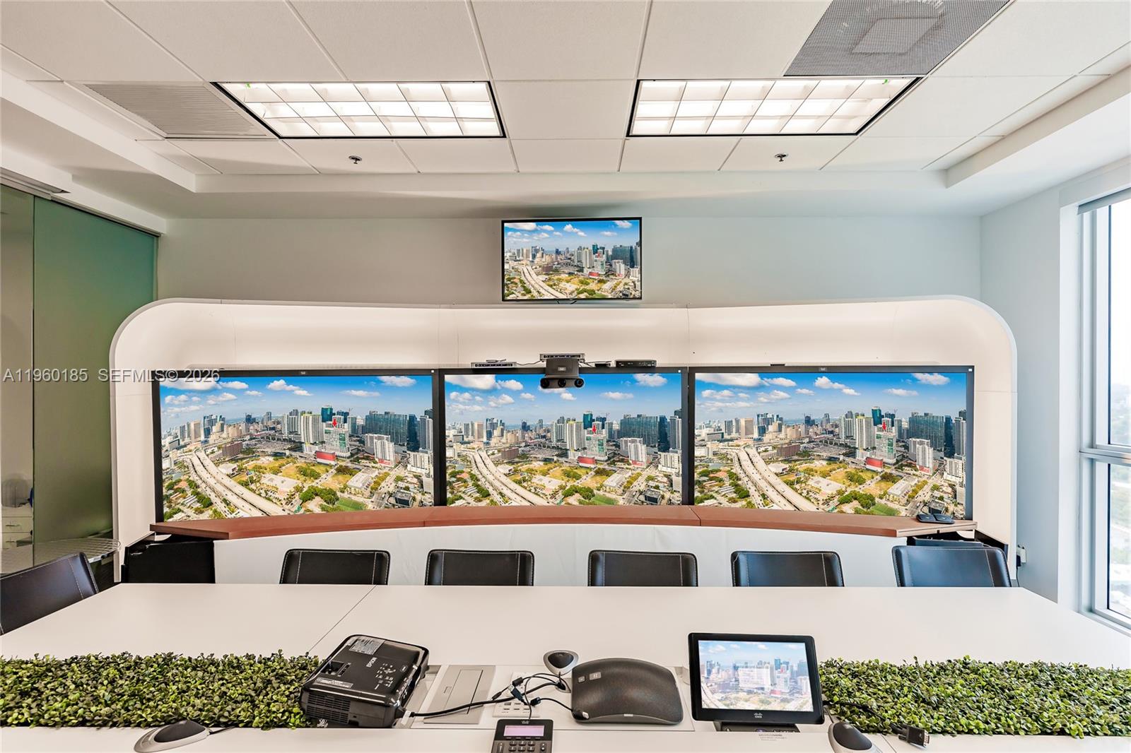 A modern conference room features a large curved desk, multiple wall-mounted monitors, and greenery accents — 175 SW 7th St # 1901, Miami FL 33130, MIAMI, FL