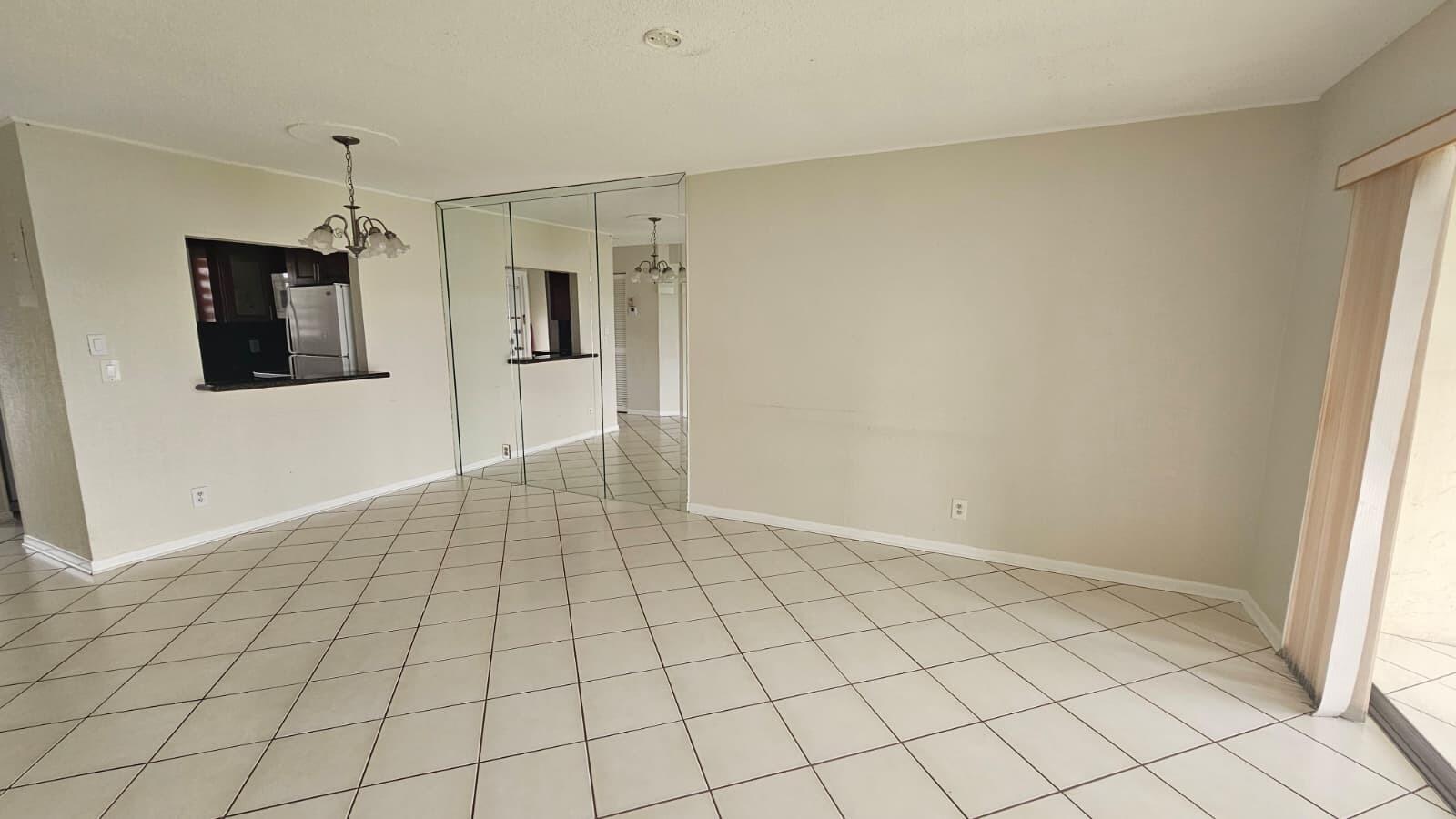 An empty room with beige tile floors, a mirrored wall, and a kitchen pass-through window — 9911 Sandalfoot Boulevard # 116, Boca Raton FL 33428, BOCA RATON, FL