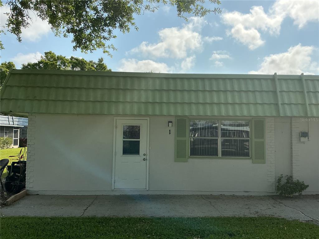 A single-story building exterior features a white door, a window with green shutters, and a green roof — 1802 BEDFORD LANE UNIT 1, SUN CITY CENTER, FL