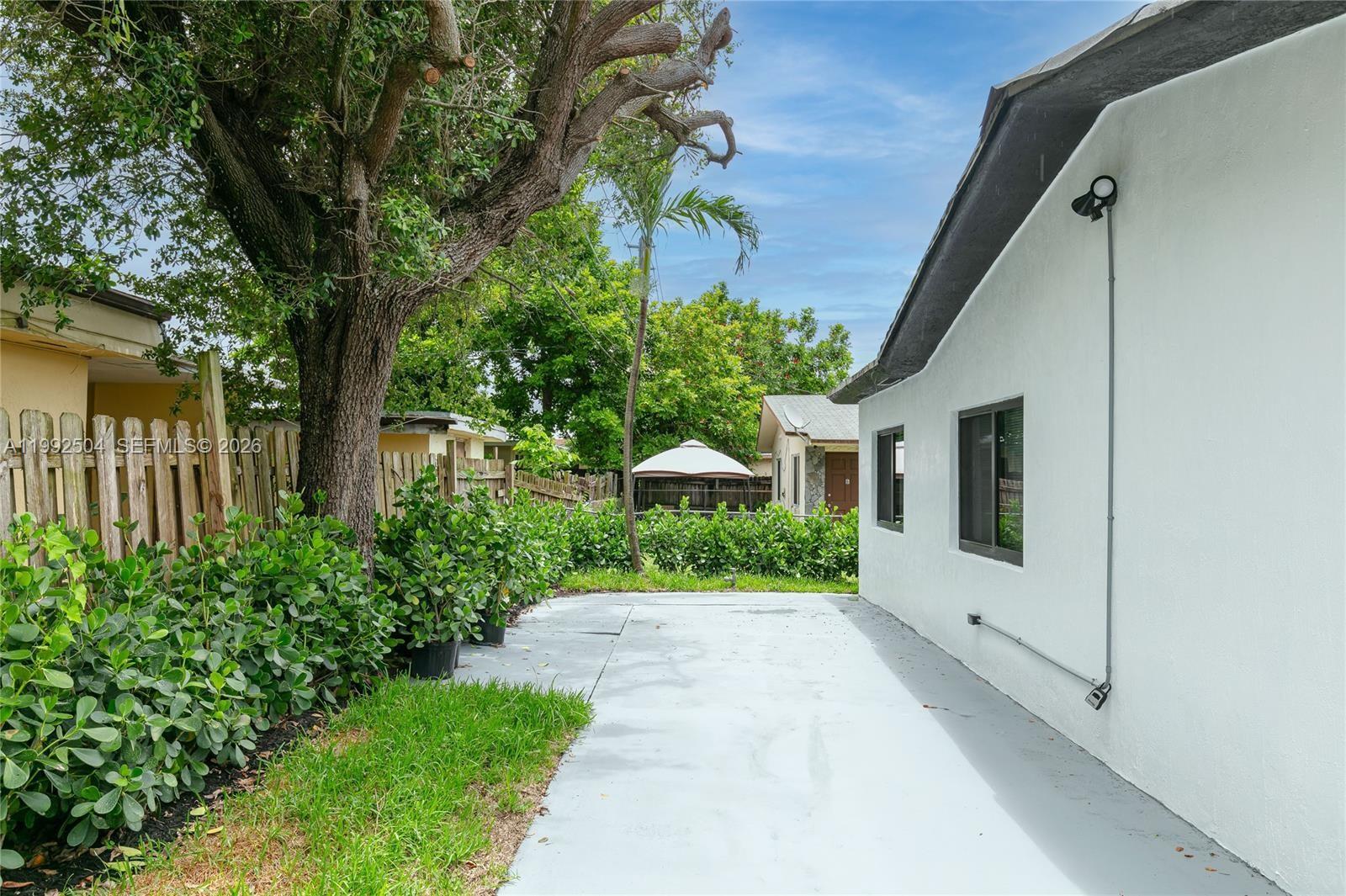 A concrete patio sits beside a white house wall under a large tree — 6125 SW 40th Ct, Miramar FL 33023, MIRAMAR, FL