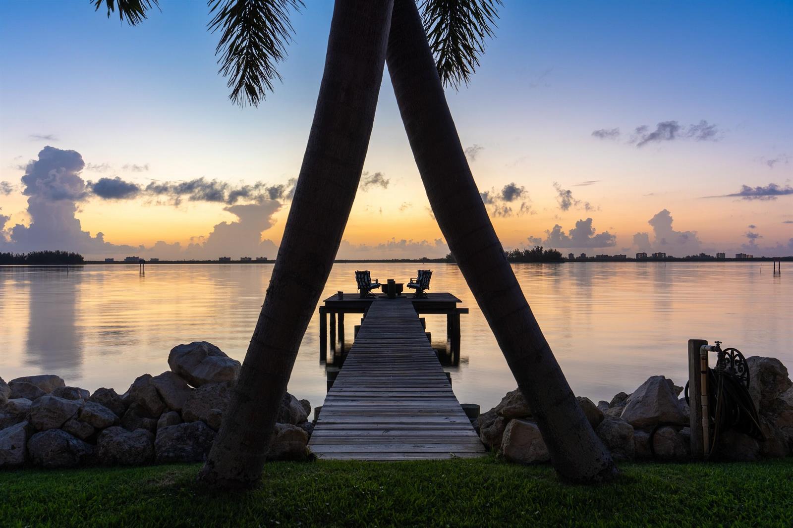 A wooden dock with two chairs extends into calm water between two palm tree trunks — FORT PIERCE, FL