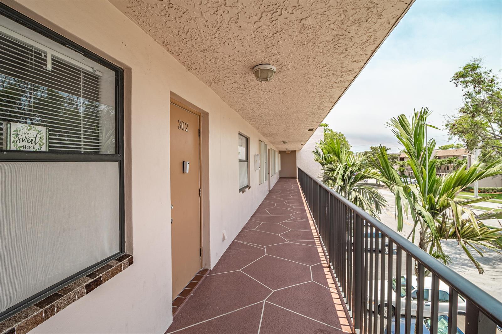 An exterior walkway with a tan door, metal railing, and palm trees in the background — 10331 Sunrise Lakes Boulevard # 302, Sunrise FL 33322, SUNRISE, FL