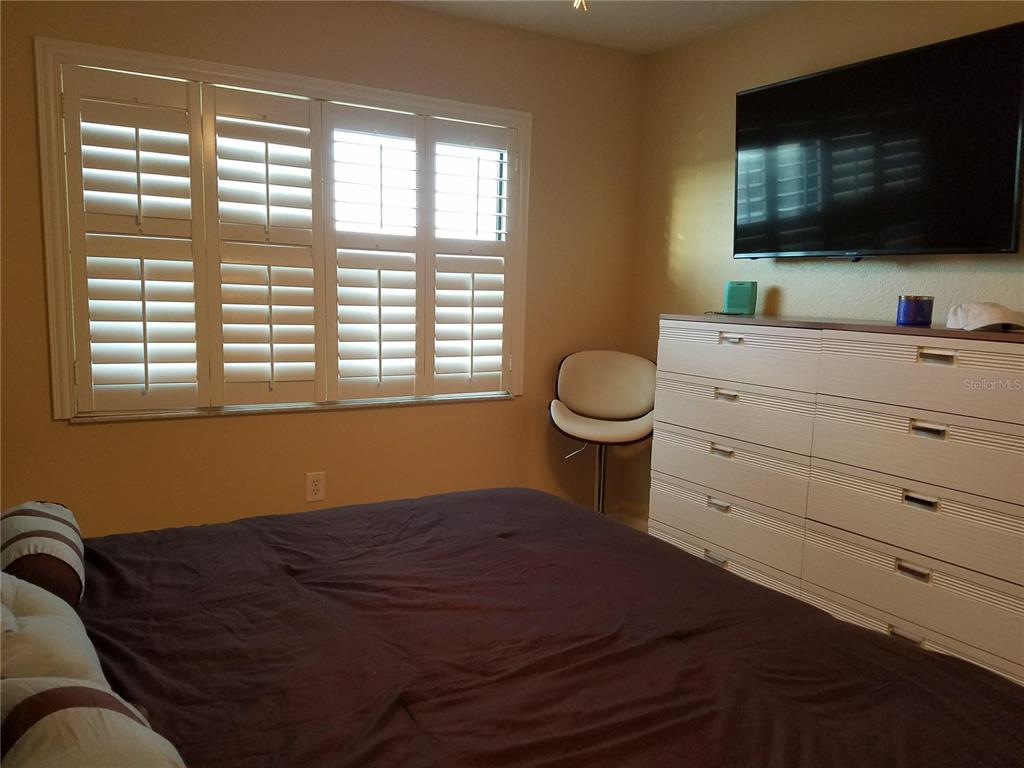 A bedroom features a bed, a white dresser, a wall-mounted television, and plantation shutters — 1571 S ATLANTIC AVENUE UNIT 211, NEW SMYRNA BEACH, FL