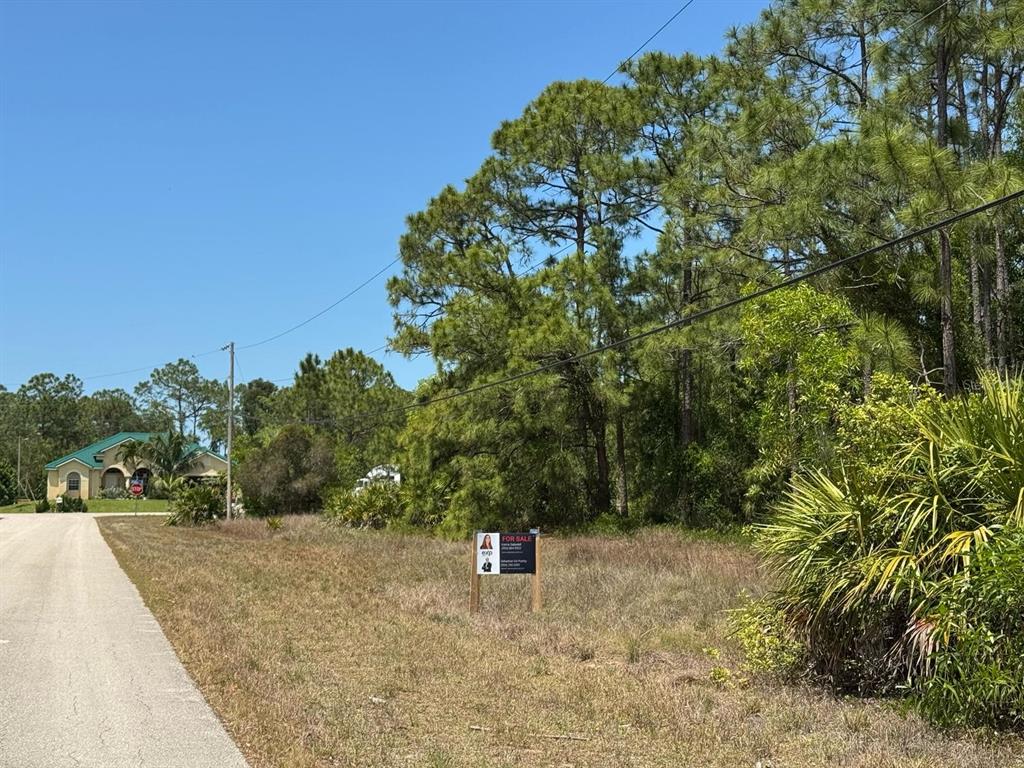 A vacant, grassy lot with a for sale sign sits beside a paved road — 513 ADDISON STREET E, LEHIGH ACRES, FL