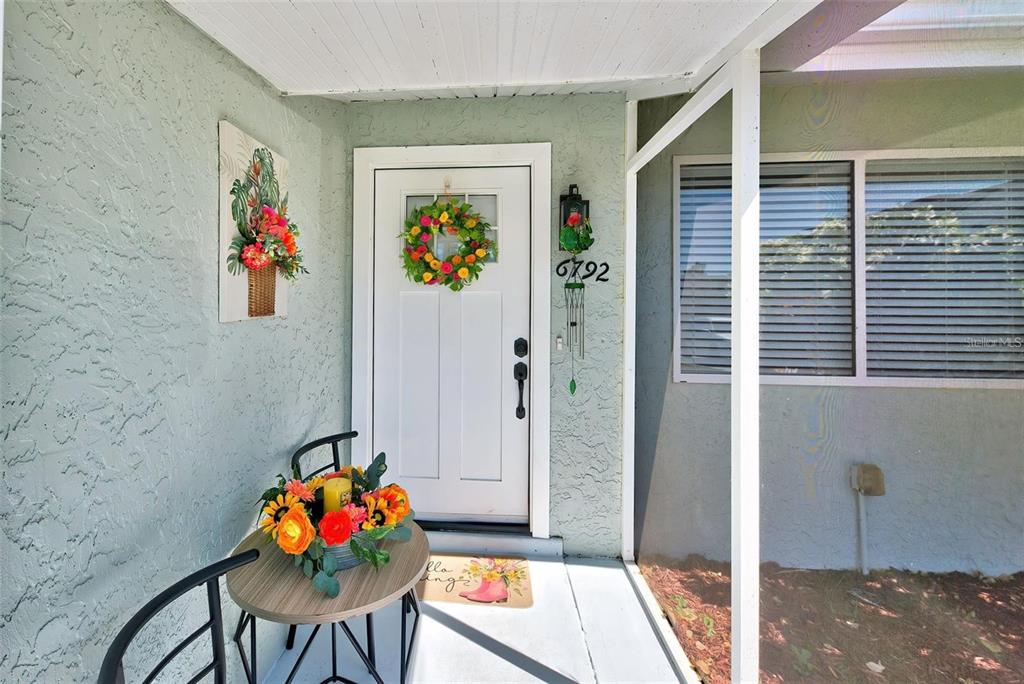 A white front door with a floral wreath sits on a covered porch with stucco walls — 6792 HIGDON ROAD, NORTH PORT, FL