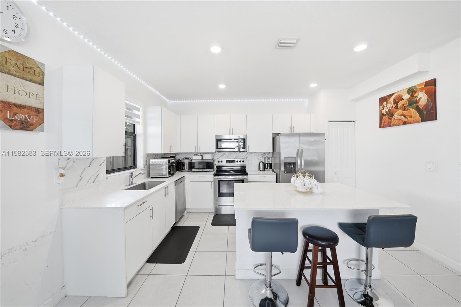 A bright white kitchen features stainless steel appliances, a central island, and tile flooring — 13091 SW 232nd Ln, Miami FL 33170, MIAMI, FL