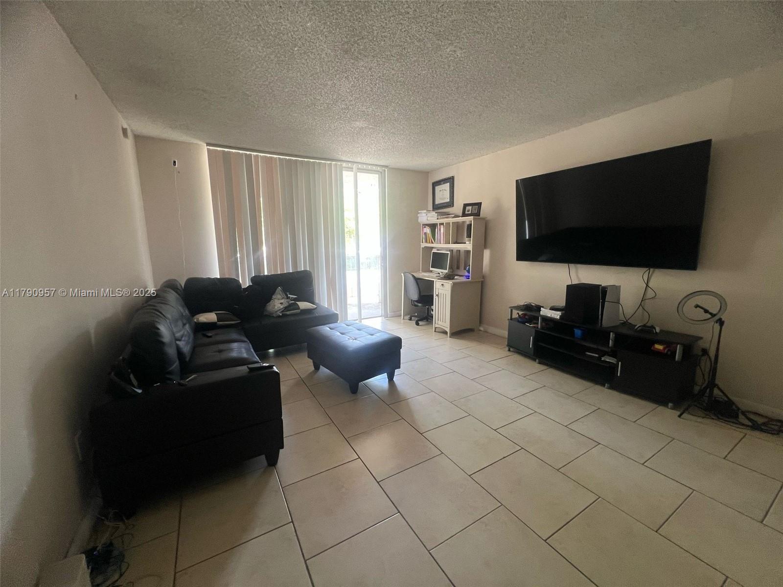 A living room features tile floors, a black sectional sofa, a wall-mounted television, and a desk — 482 NW 165th St Rd # A203, Miami FL 33169, MIAMI, FL