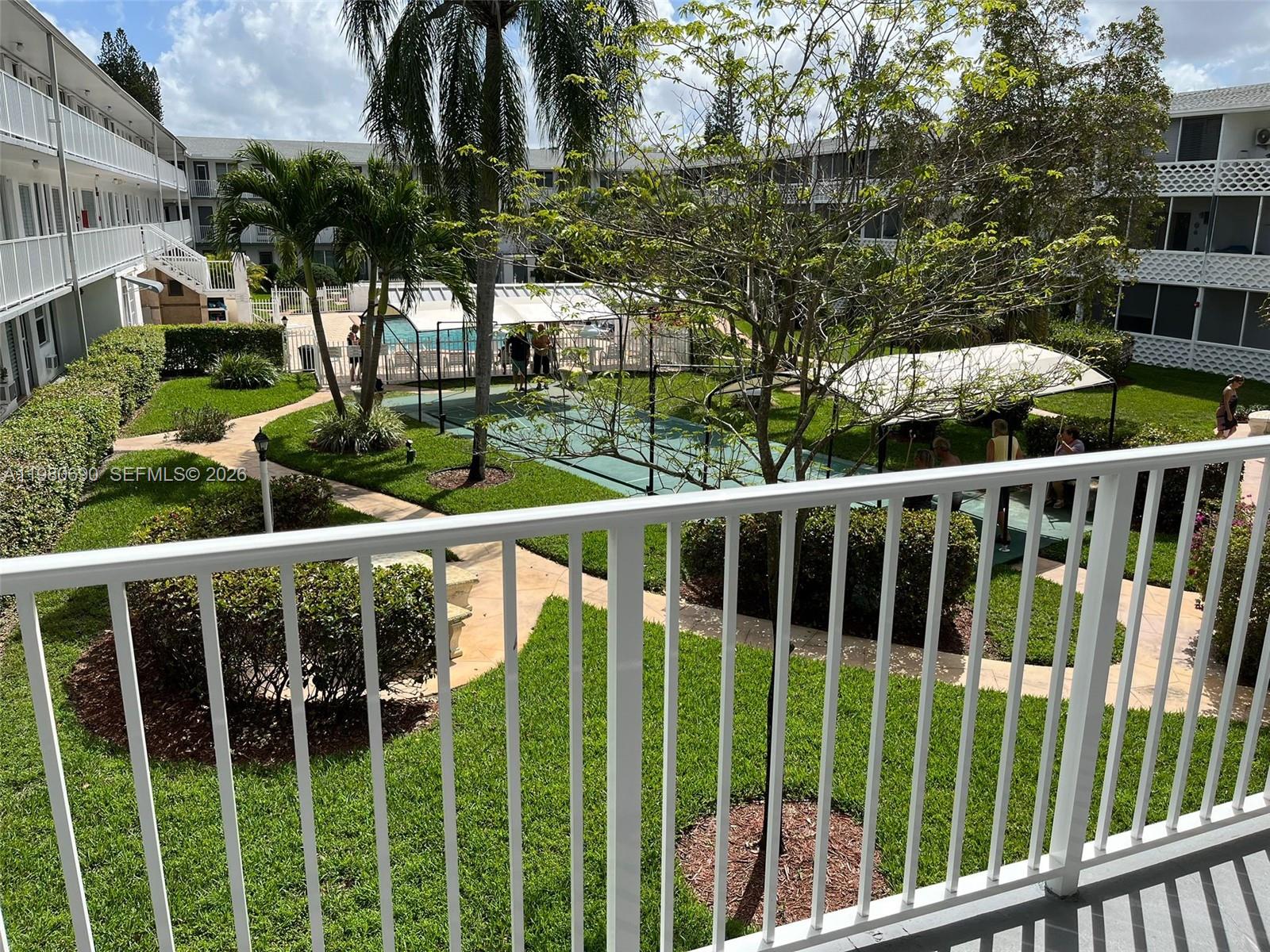 A balcony view overlooks a courtyard with a swimming pool, tennis court, and landscaping — 15610 NE 6th Ave # 19C, Miami FL 33162, MIAMI, FL