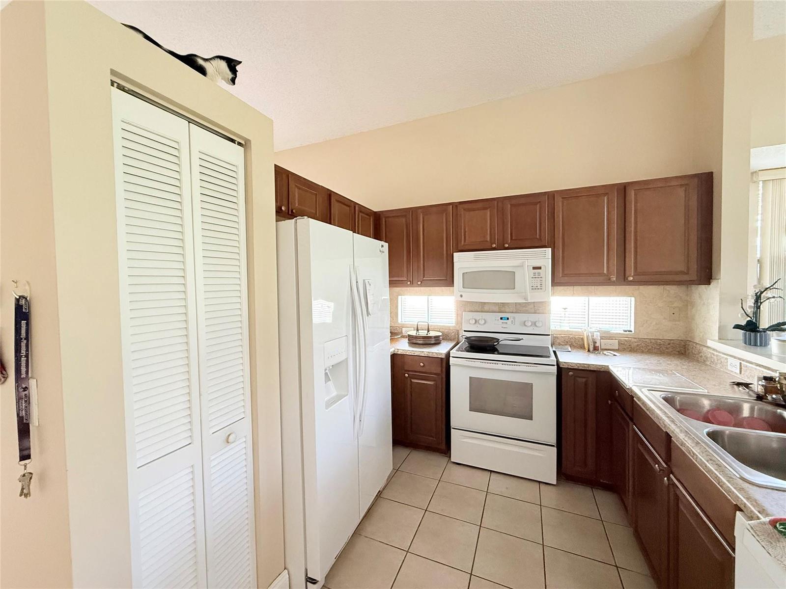 A kitchen with brown cabinets, white appliances, tile flooring, and a cat perched above the pantry — 4965 SW 153rd Avenue, Davie FL 33331, DAVIE, FL