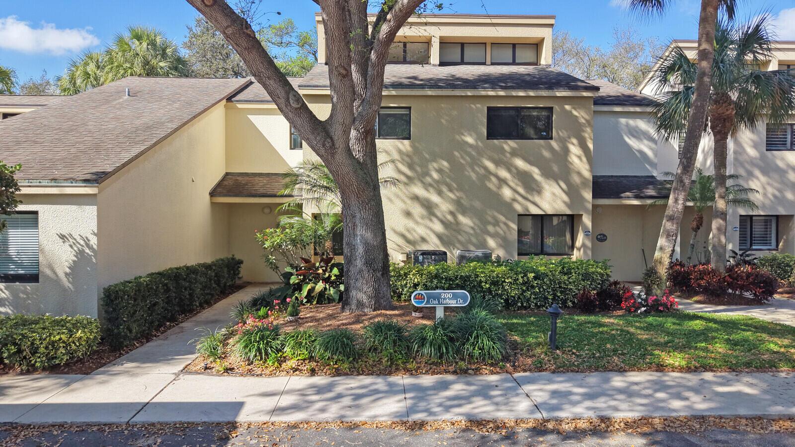 A two-story townhouse exterior features tan stucco walls, a shingled roof, and a large tree — 215 Oak Harbour Drive, Juno Beach FL 33408, JUNO BEACH, FL