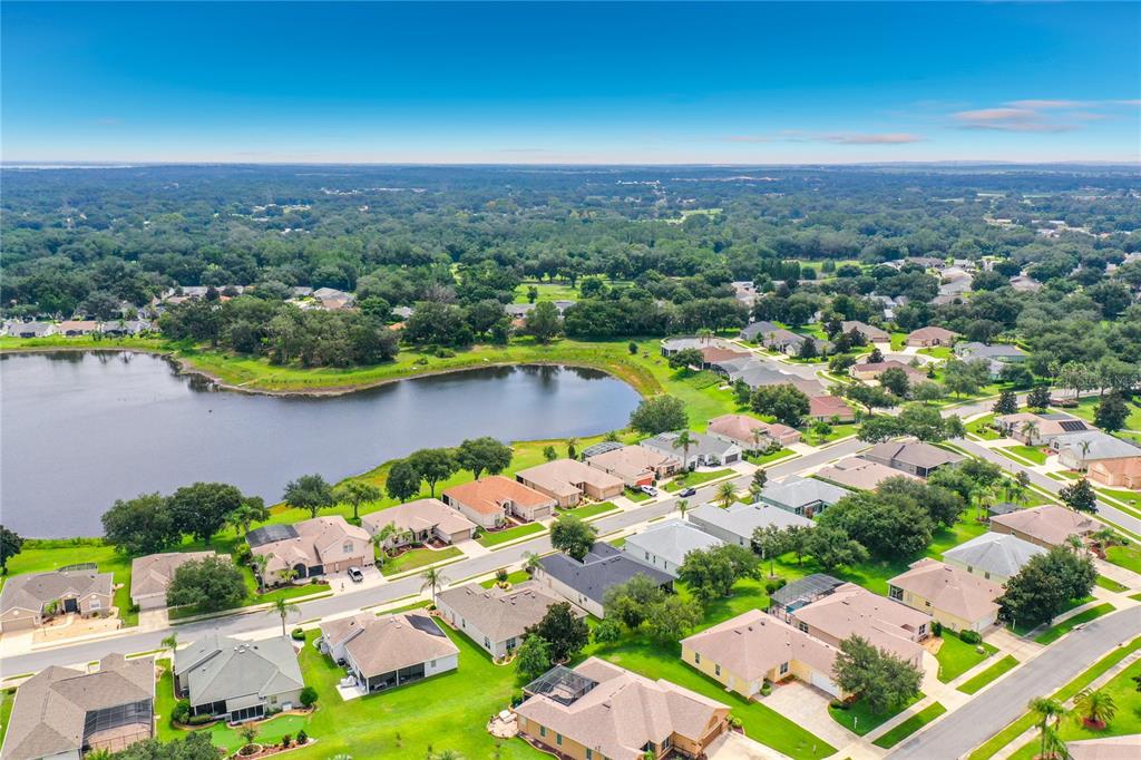 An aerial view shows suburban houses surrounding a lake with dense trees in the background — 25524 LAUREL VALLEY ROAD, LEESBURG, FL