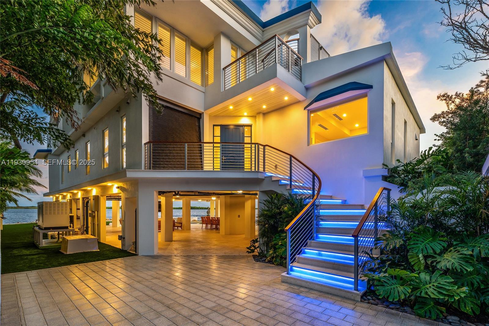A modern multi-story waterfront home features an elevated entrance with illuminated stairs and paved driveway — 180 Pompano Dr, Key Largo FL 33037, KEY LARGO, FL
